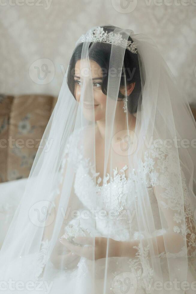 portrait. le brunette la mariée est permanent dans une élégant couronne et blanc robe, enveloppé dans une voile, à la recherche vers le bas et reniflement le du marié boutonnière. magnifique maquillage et cheveux. volumineux voile. photo