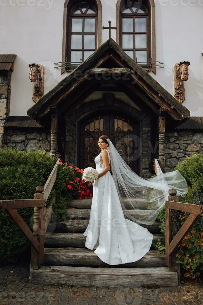 romantique toute la longueur portrait de une cheveux bruns femme dans une blanc robe avec une bouquet permanent près le en bois porte de le église entrée photo