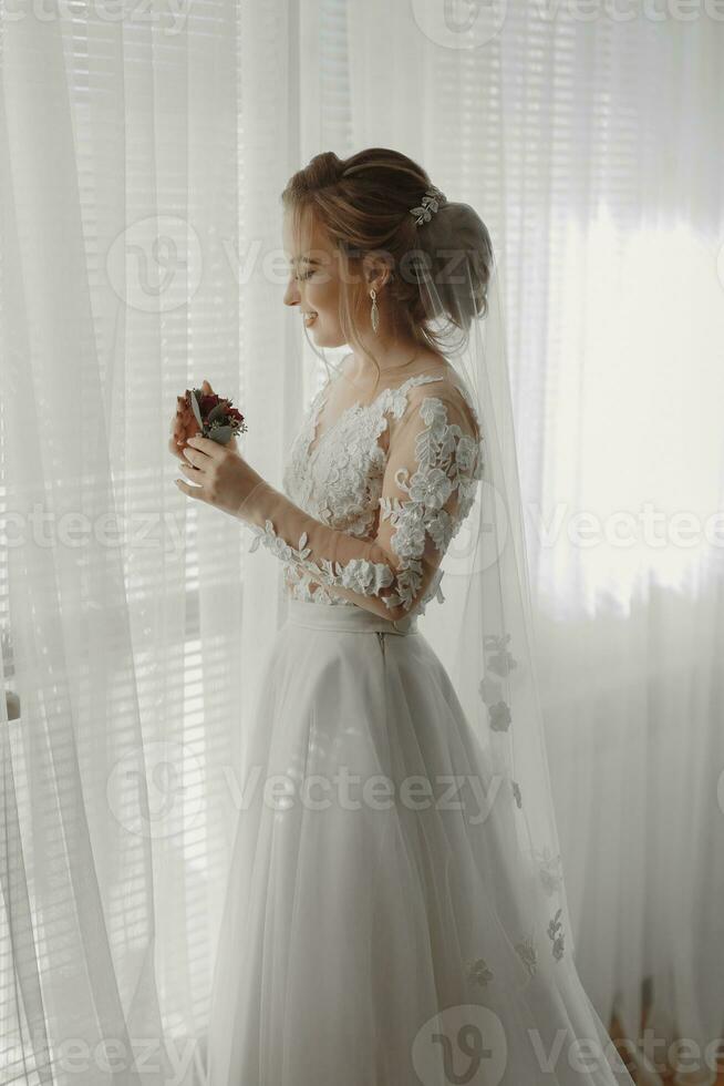 préparation pour le mariage. magnifique Jeune la mariée dans blanc mariage robe à l'intérieur. luxe modèle regards à mariage chaussures, à Accueil dans studio pièce avec gros la fenêtre. le fille spectacles photo