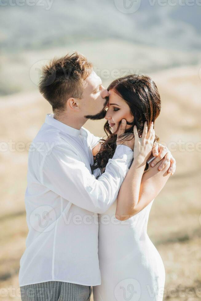 une élégant modèle couple dans le montagnes dans le été. une Jeune garçon et une fille dans une blanc soie robe sont en marchant sur le pente contre le Contexte de le forêt et Montagne pics. garçon baisers une fille photo