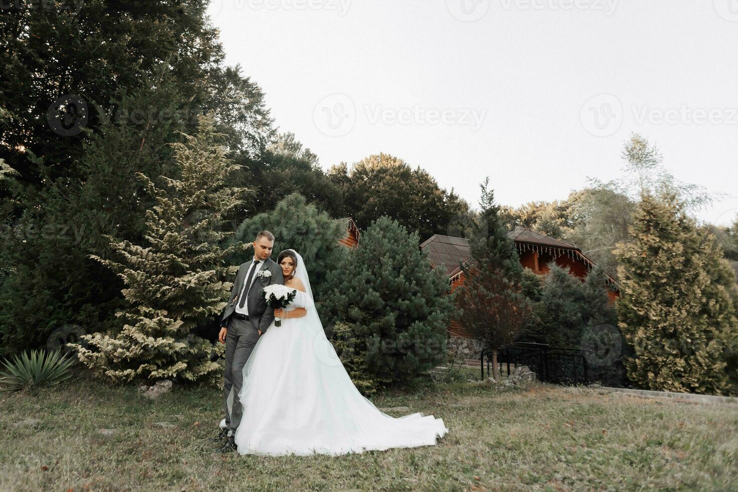 portrait de le la mariée et jeune marié dans la nature. le jeune marié est dans le premier plan, flou, le la mariée des stands derrière lui dans une blanc volumineux robe, en portant une bouquet et à la recherche bas. élégant jeune marié photo