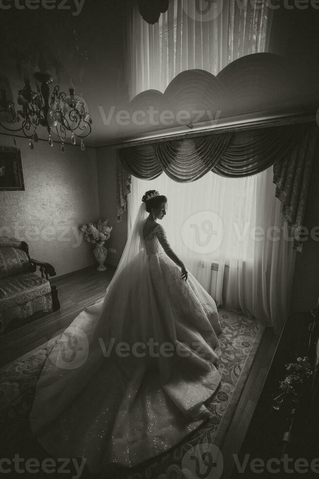 le la mariée dans une blanc mariage robe. content magnifique Jeune femme dans blanc traditionnel mariage robe avec longue train dans sa chambre. grand les fenêtres et lumière des murs. noir et blanc photo