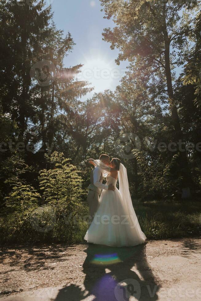 portrait de une magnifique couple dans l'amour sur votre mariage journée. une marcher dans le parc dans le ensoleillement et verdure. incroyable baisers et câlins de le la mariée et jeune marié avec une bouquet photo