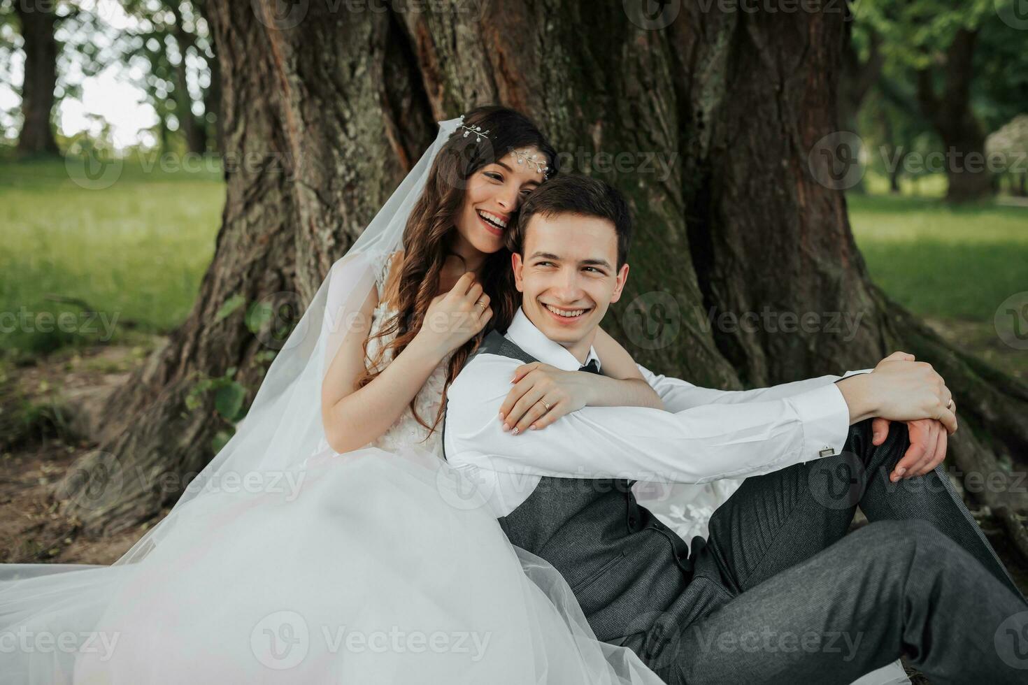 une magnifique la mariée avec longue frisé cheveux dans une élégant robe câlins le marié, sourires, à la recherche dans le lentille en dessous de une gros arbre. portrait de le la mariée et jeune marié. printemps mariage. Naturel maquillage photo