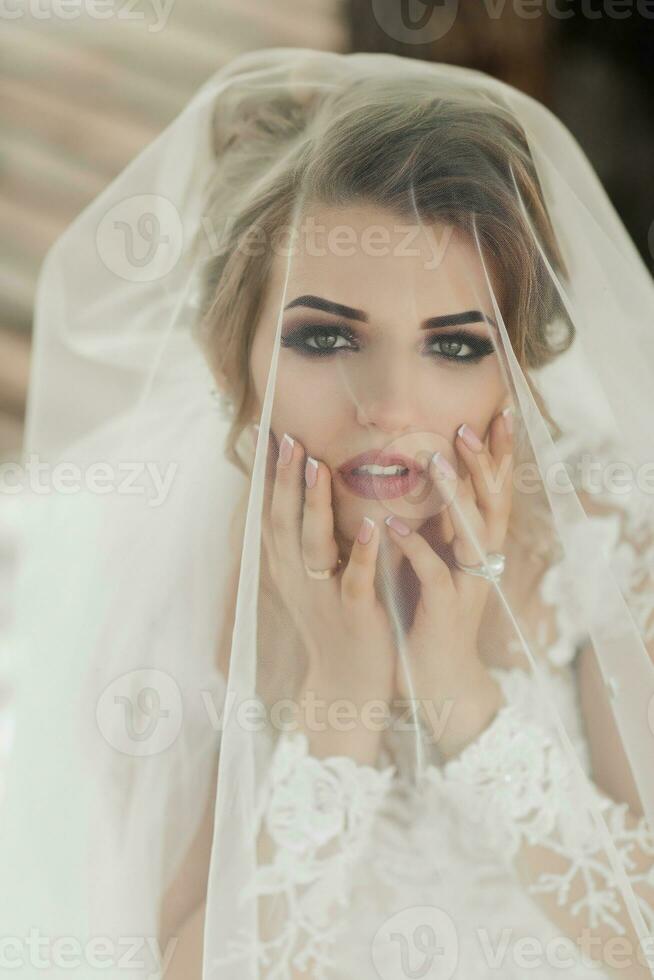 frisé blond la mariée dans une blanc robe, couvert avec une voile, pose pour le caméra avec une bouquet de des roses. portrait de le la mariée. magnifique maquillage et cheveux. mariage dans la nature photo