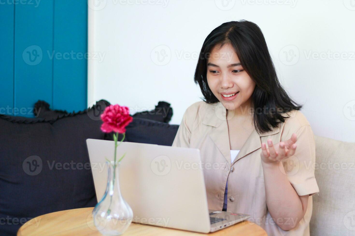 souriant asiatique femme travail sur portable avec élevage paumes séance sur canapé portant décontractée vêtements, en présentant geste à l'intérieur photo