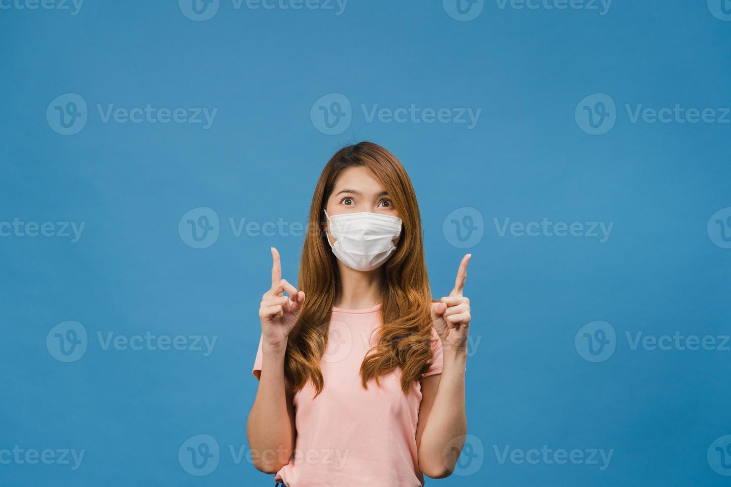 une jeune fille asiatique porte un masque médical montre quelque chose dans un espace vide vêtu d'un tissu décontracté et regarde la caméra isolée sur fond bleu. distanciation sociale, quarantaine pour le virus corona. photo