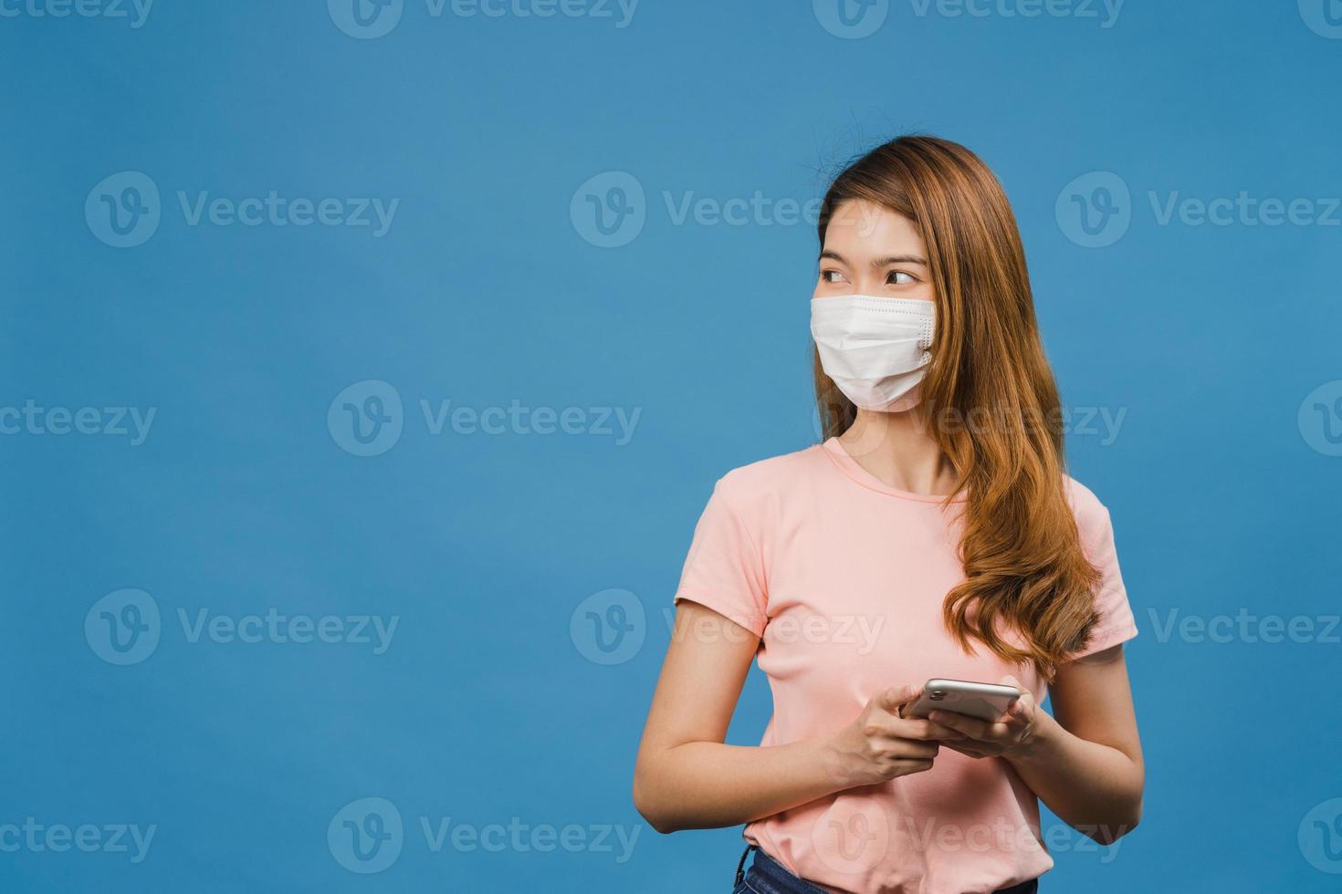 jeune fille asiatique portant un masque médical utilisant un téléphone portable avec des vêtements décontractés isolés sur fond bleu. auto-isolement, distanciation sociale, quarantaine pour la prévention du virus corona. photo