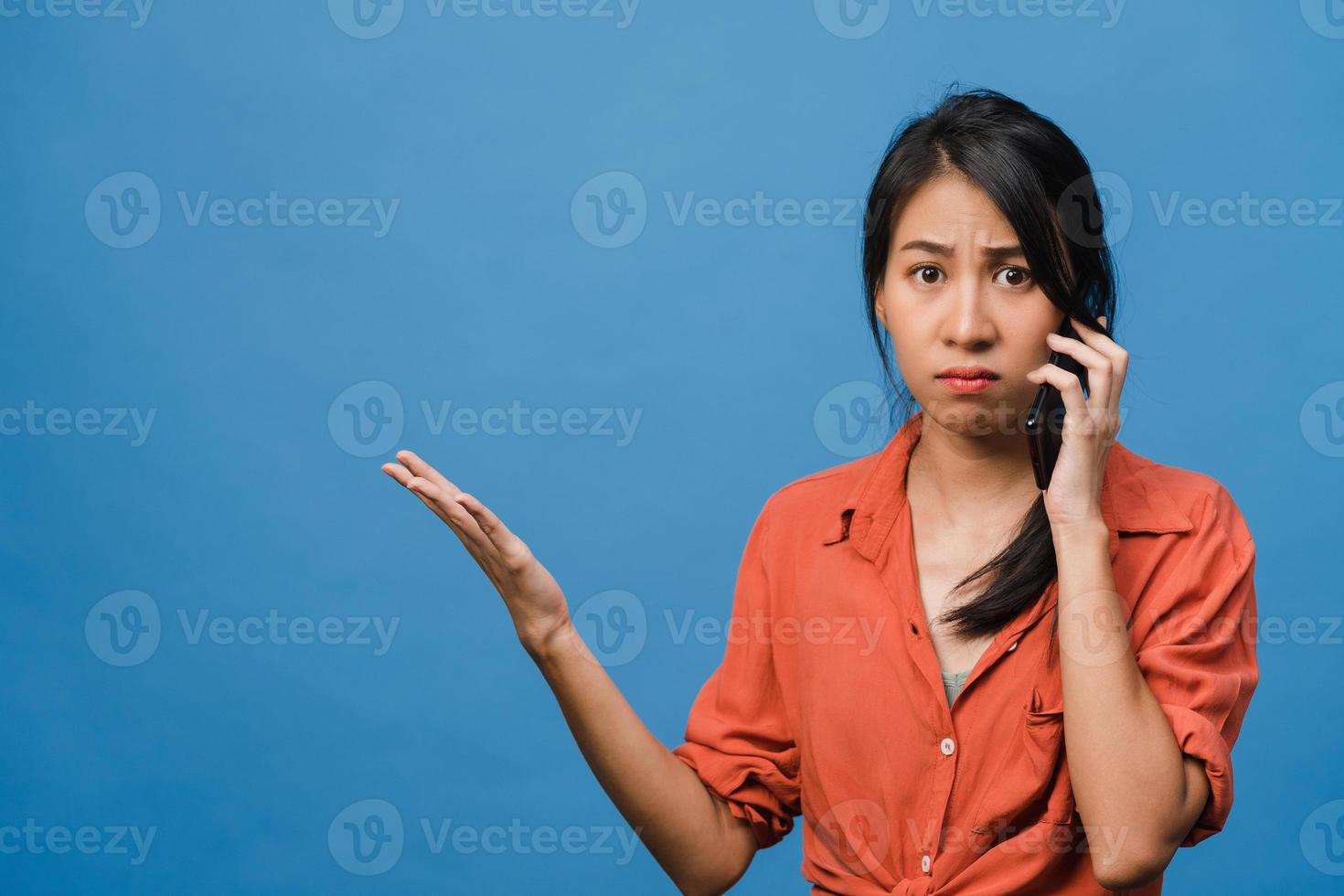 une jeune femme asiatique parle par téléphone avec une expression négative, des cris excités, des cris émotionnels en colère dans un tissu décontracté et se tient isolée sur fond bleu avec un espace de copie vierge. concept d'expression faciale. photo