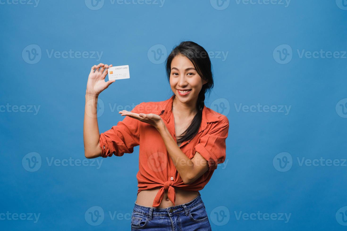 jeune femme asiatique montre une carte bancaire de crédit avec une expression positive, sourit largement, vêtue de vêtements décontractés, se sentant heureuse et isolée sur fond bleu. concept d'expression faciale. photo