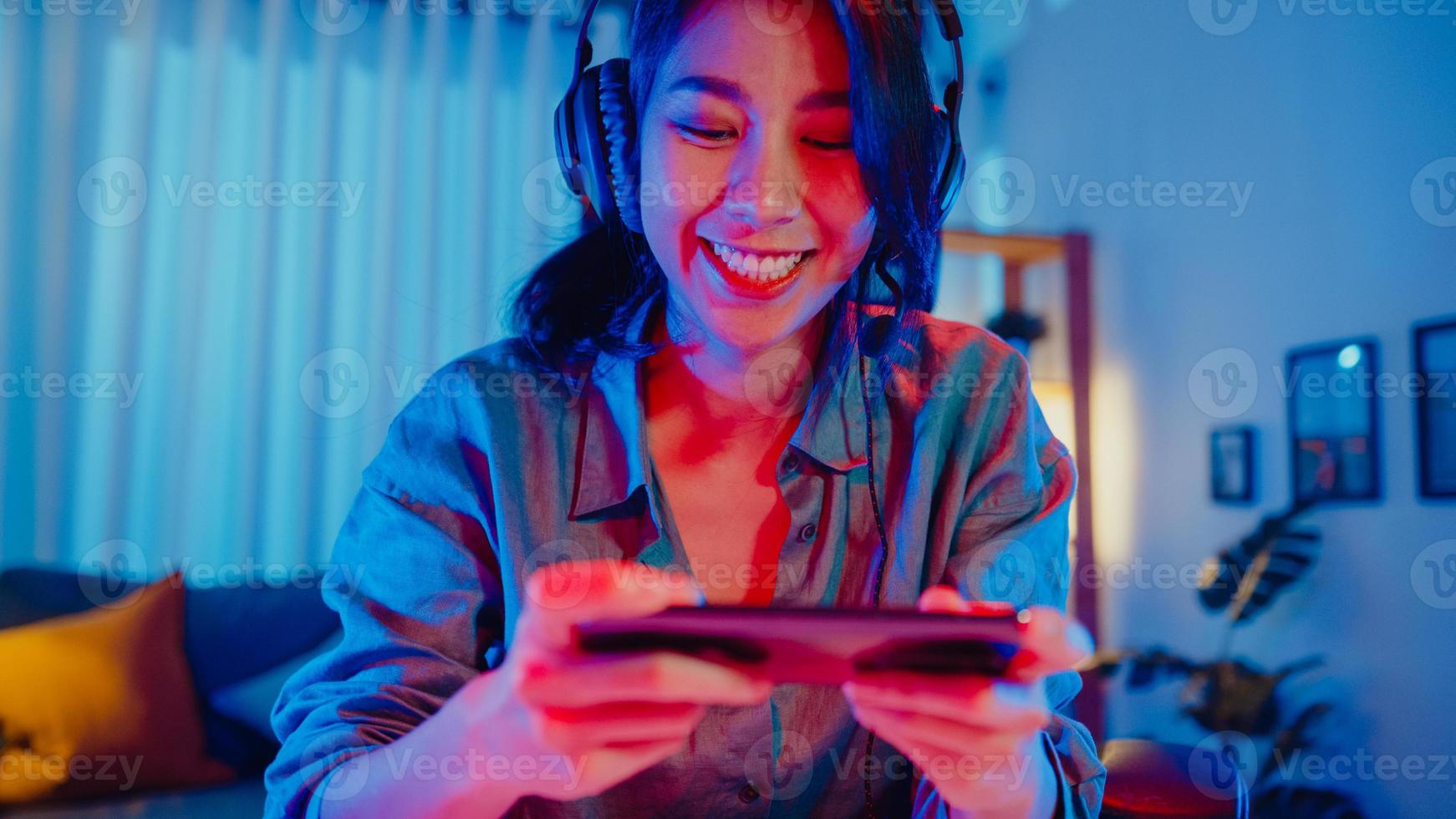 Happy asia girl gamer porter un concours de casque jouer à un jeu vidéo en ligne avec des néons colorés pour smartphone dans le salon à la maison. jeu de streaming esport en ligne, concept d'activité de quarantaine à domicile. photo