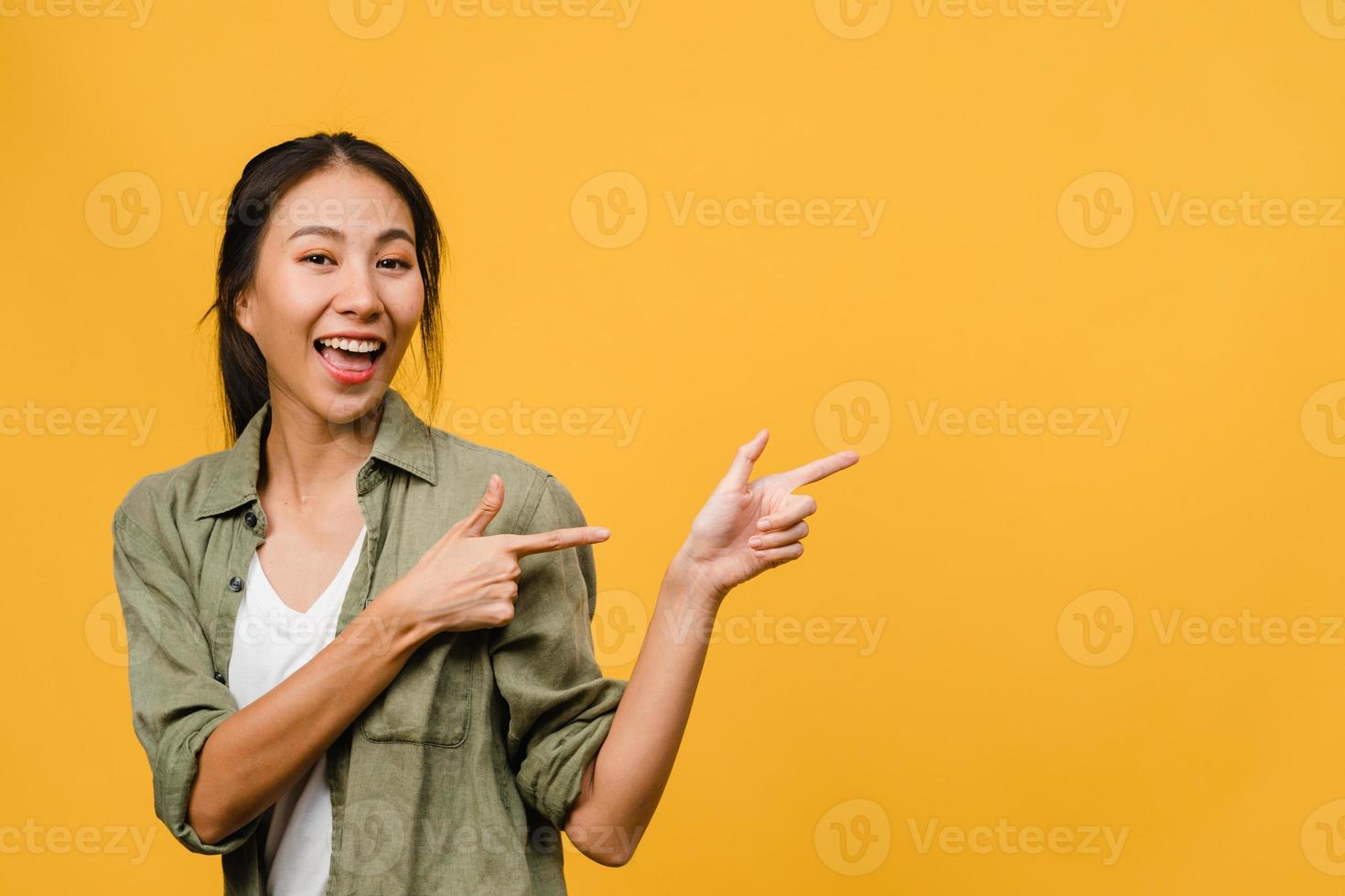 portrait d'une jeune femme asiatique souriante avec une expression joyeuse, montre quelque chose d'étonnant dans un espace vide dans un tissu décontracté et regardant la caméra isolée sur fond jaune. concept d'expression faciale. photo