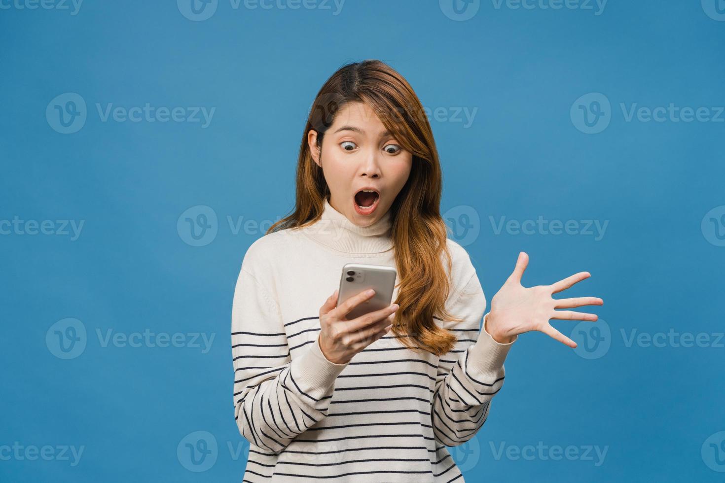 surpris une jeune femme asiatique utilisant un téléphone portable avec une expression positive, sourit largement, vêtue de vêtements décontractés et debout isolée sur fond bleu. heureuse adorable femme heureuse se réjouit du succès. photo