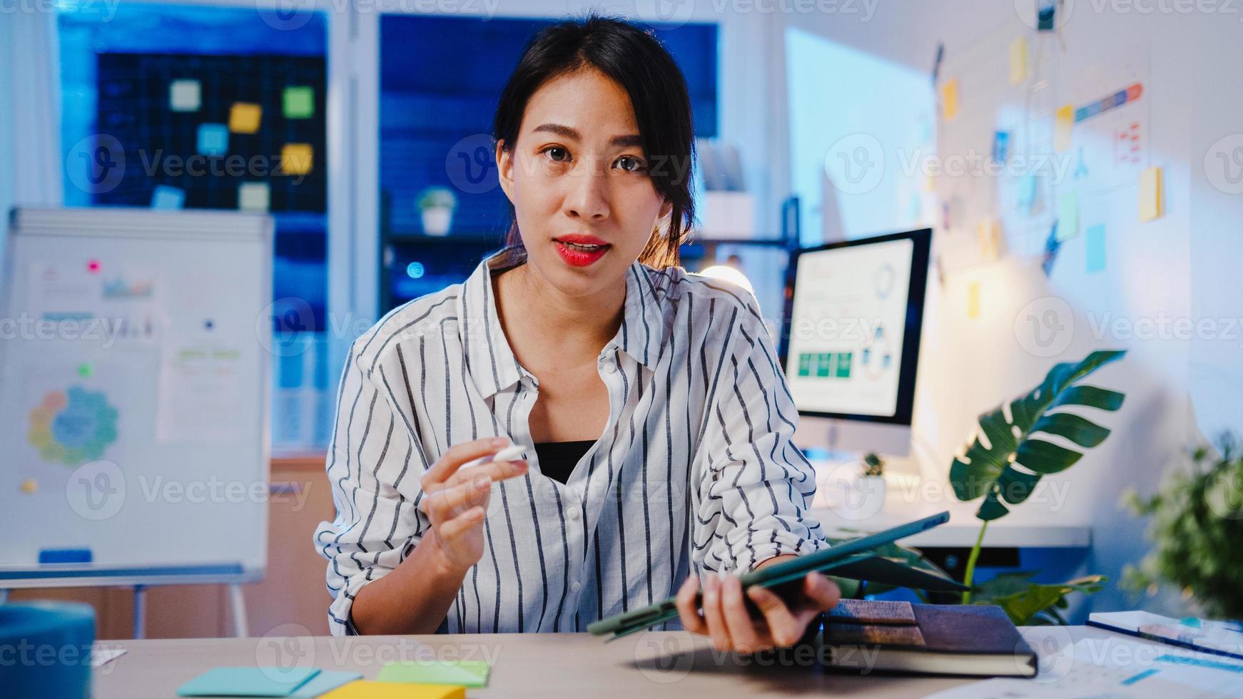 femme d'affaires asiatique distanciation sociale dans une nouvelle norme pour la prévention des virus en regardant la présentation de la caméra à un collègue au sujet du plan en appel vidéo pendant le travail de nuit au bureau. mode de vie après le coronavirus. photo