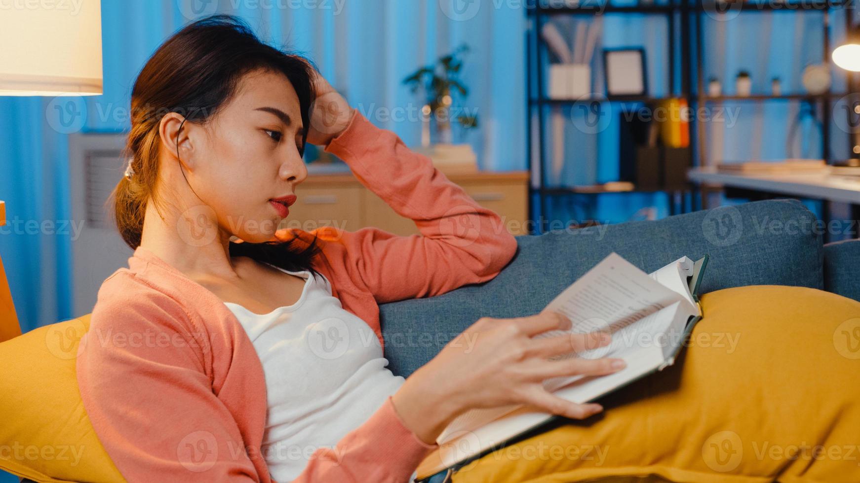 la nuit, une belle dame asiatique a lu un livre avec bonheur dans un salon paisible sur un canapé. éducation à domicile, rester à la maison, activité d'auto-quarantaine, activité amusante pour la quarantaine covid ou coronavirus. photo
