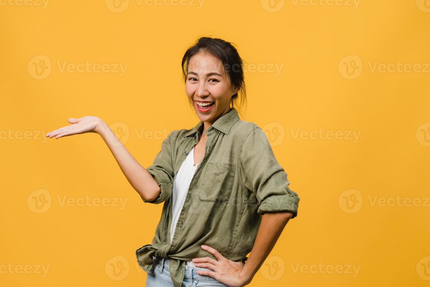 portrait d'une jeune femme asiatique souriante avec une expression joyeuse, montre quelque chose d'étonnant dans un espace vide dans un tissu décontracté et regardant la caméra isolée sur fond jaune. concept d'expression faciale. photo