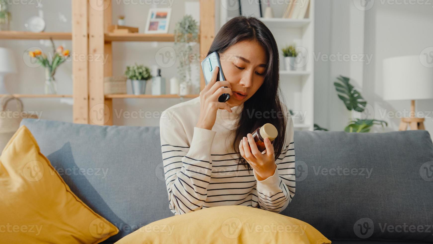 une jeune femme asiatique malade tient un médicament assis sur un canapé utilise un appel de smartphone pour consulter un médecin à la maison. la fille prend des médicaments après l'ordonnance du médecin, mise en quarantaine à la maison, concept de coronavirus de distanciation sociale. photo