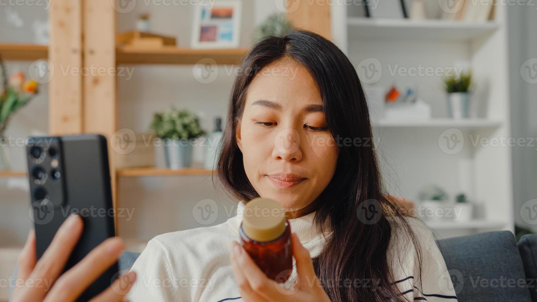 jeune femme asiatique malade tenir un médicament assis sur un canapé appel vidéo avec téléphone consulter un médecin à la maison. la fille prend des médicaments après l'ordonnance du médecin, mise en quarantaine à la maison, concept de coronavirus de distanciation sociale. photo