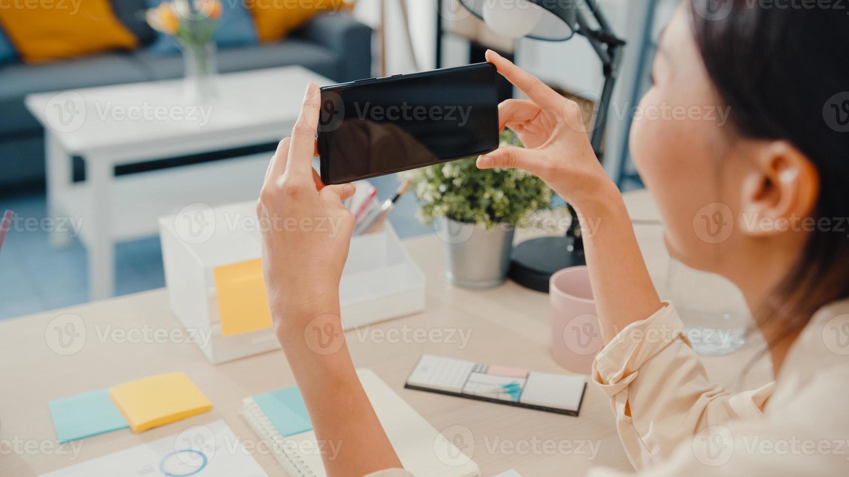 une jeune femme asiatique utilise un téléphone intelligent avec un écran noir vierge pour le texte publicitaire tout en travaillant à domicile dans le salon. technologie de clé de chrominance, concept de design marketing. photo