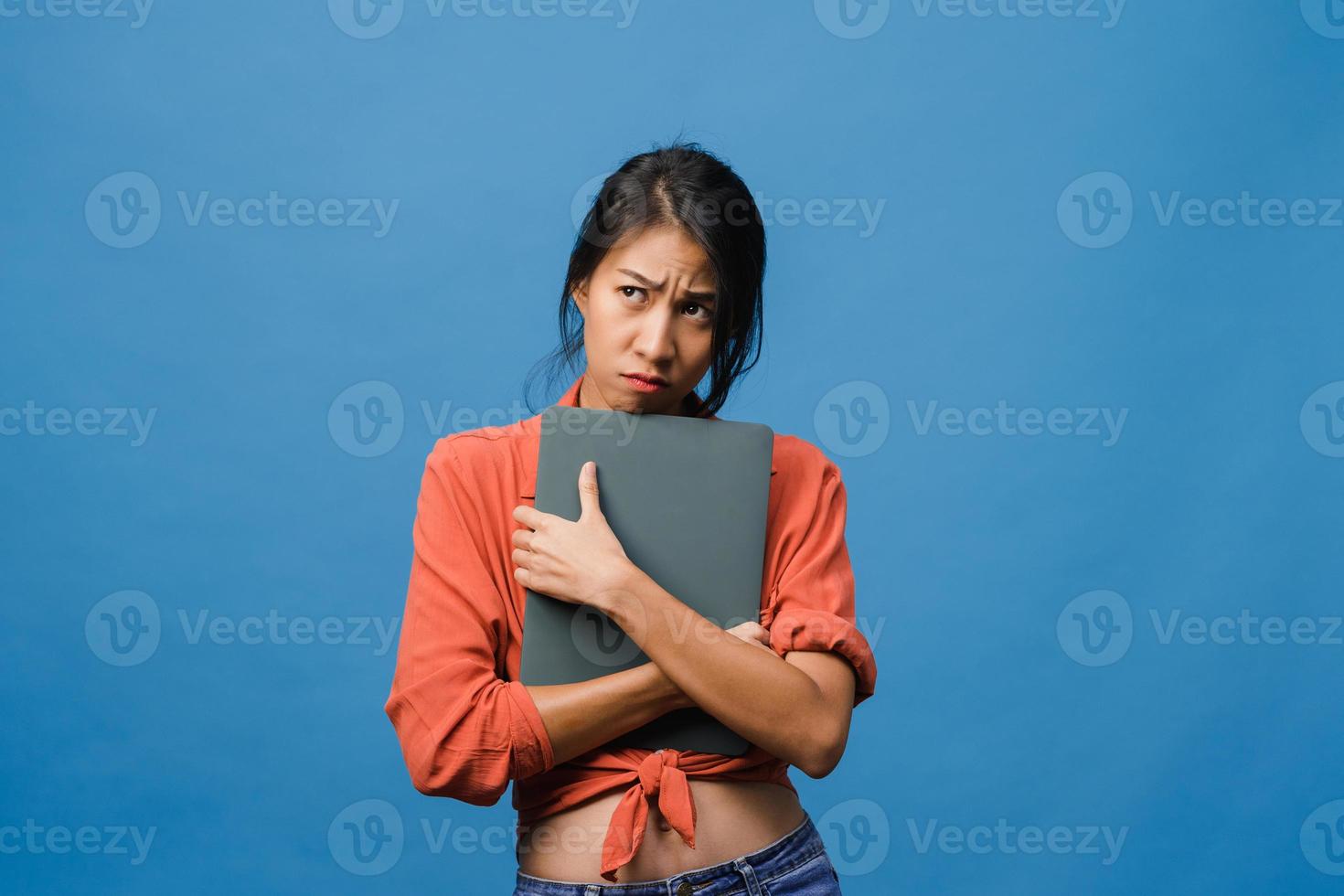une jeune femme asiatique tient un ordinateur portable avec une expression négative, des cris excités, des cris émotionnels en colère dans un tissu décontracté et se tient isolée sur fond bleu avec un espace de copie vierge. concept d'expression faciale. photo
