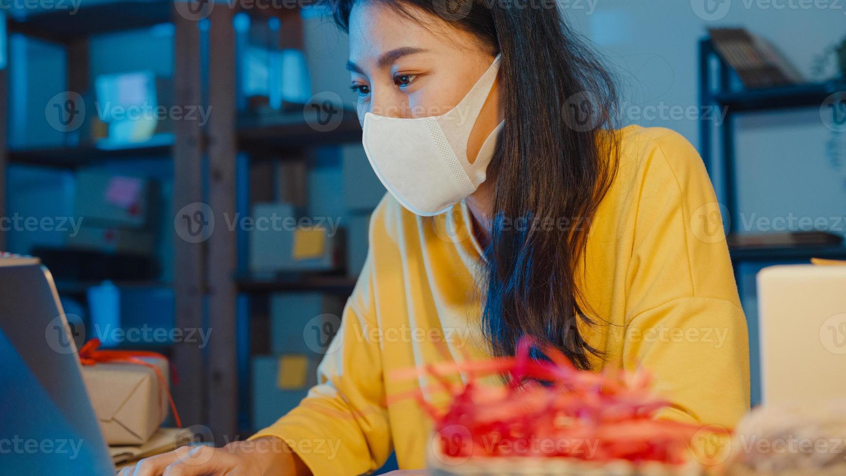 une jeune femme d'affaires asiatique porte un masque facial, vérifie le bon de commande et répond au chat de la boîte de réception du client sur un ordinateur portable au bureau à domicile la nuit. propriétaire de petite entreprise, livraison sur le marché en ligne, concept de style de vie indépendant. photo