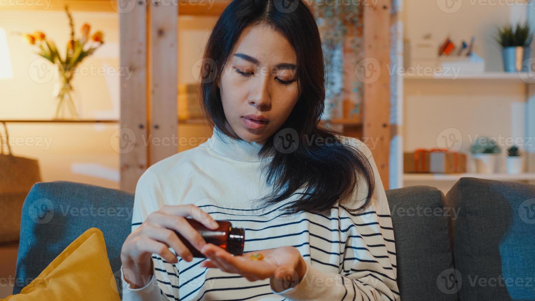 Une jeune femme asiatique malade tenant une pilule regarde la médecine s'asseoir sur un canapé à la maison la nuit. fille prenant des médicaments après l'ordonnance du médecin, mise en quarantaine à la maison, concept de soins de santé de distanciation sociale du coronavirus. photo