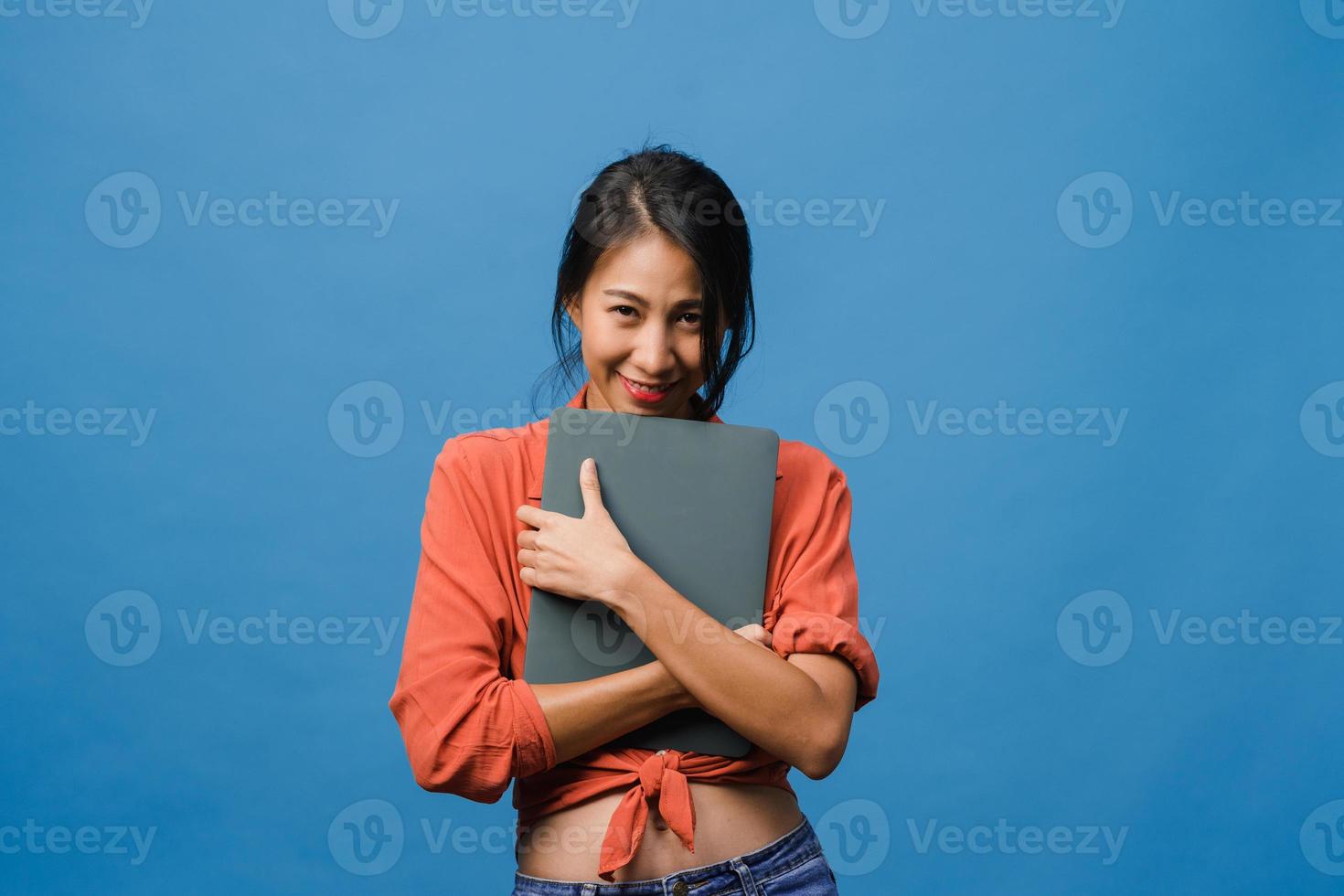 une jeune femme asiatique tient un ordinateur portable avec une expression positive, sourit largement, vêtue de vêtements décontractés, se sentant heureuse et se tient isolée sur fond bleu. heureuse adorable femme heureuse se réjouit du succès. photo