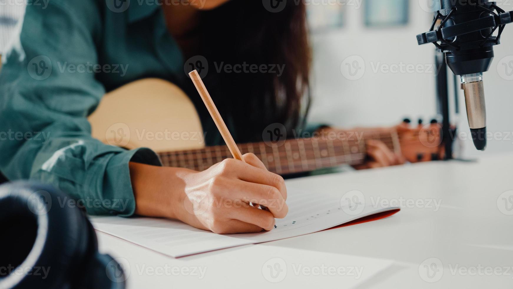 Une femme asiatique heureuse, une auteure-compositrice, joue de la guitare acoustique, écoute une chanson à partir d'un smartphone, pense et écris des paroles de chansons en papier assis dans le salon du home studio. production musicale à la maison concept. photo
