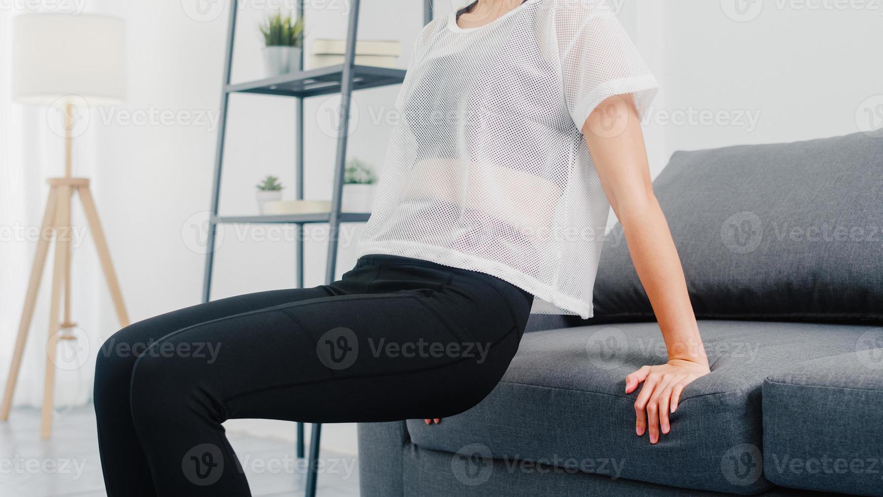 jeune femme coréenne en vêtements de sport faisant de l'exercice en faisant des trempettes triceps s'appuyant sur un canapé dans le salon à la maison. distance sociale, isolement pendant le virus. exercices pour le bas du corps. photo