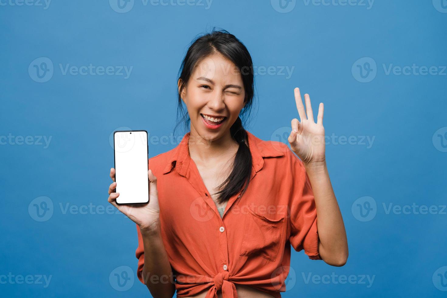 une jeune femme asiatique montre un écran de smartphone vide avec une expression positive, sourit largement, vêtue de vêtements décontractés, se sentant heureuse sur fond bleu. téléphone portable avec écran blanc en main féminine. photo