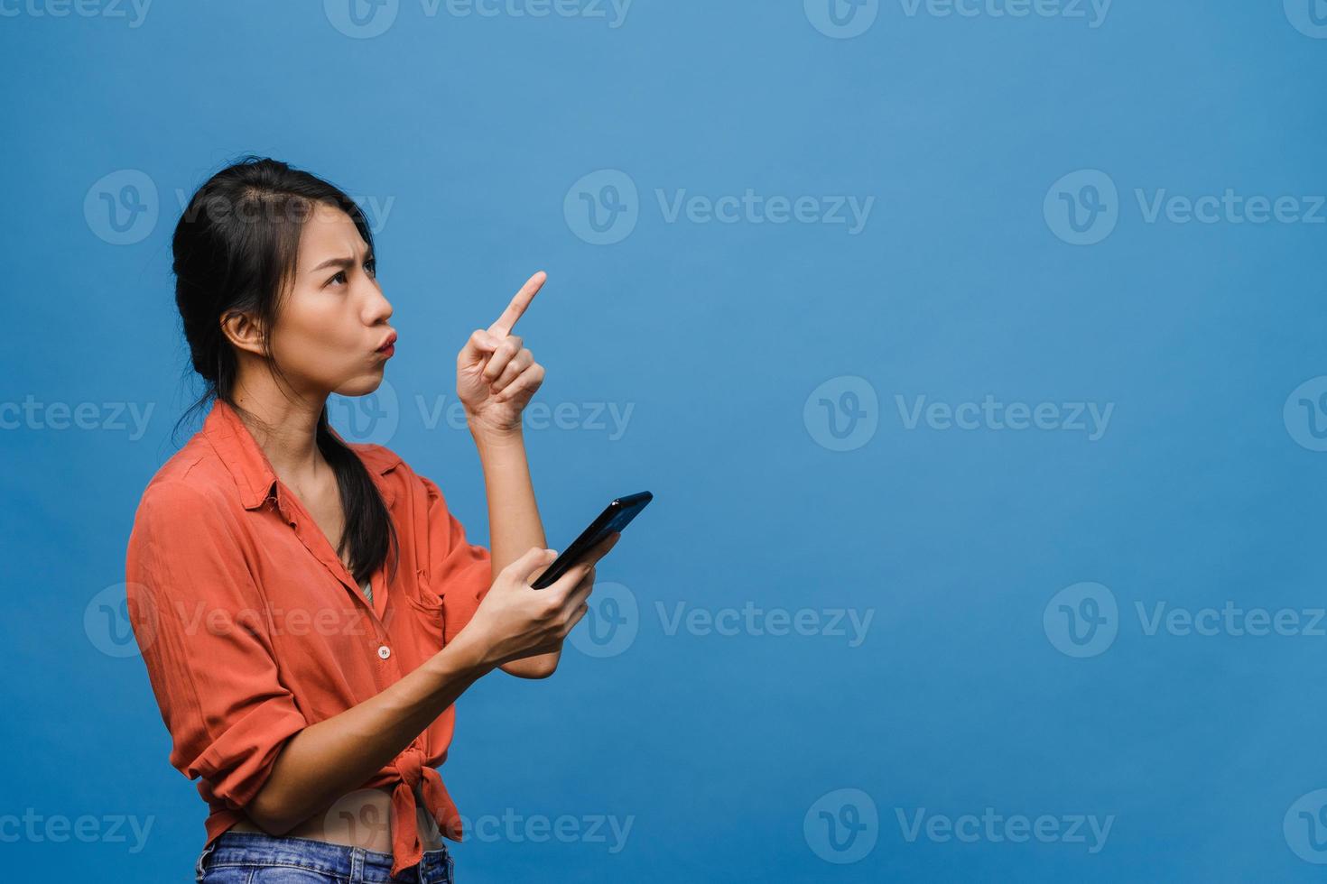 portrait d'une jeune femme asiatique utilisant un téléphone portable avec une expression joyeuse, montre quelque chose d'étonnant dans un espace vide dans des vêtements décontractés et se tient isolé sur fond bleu. concept d'expression faciale. photo