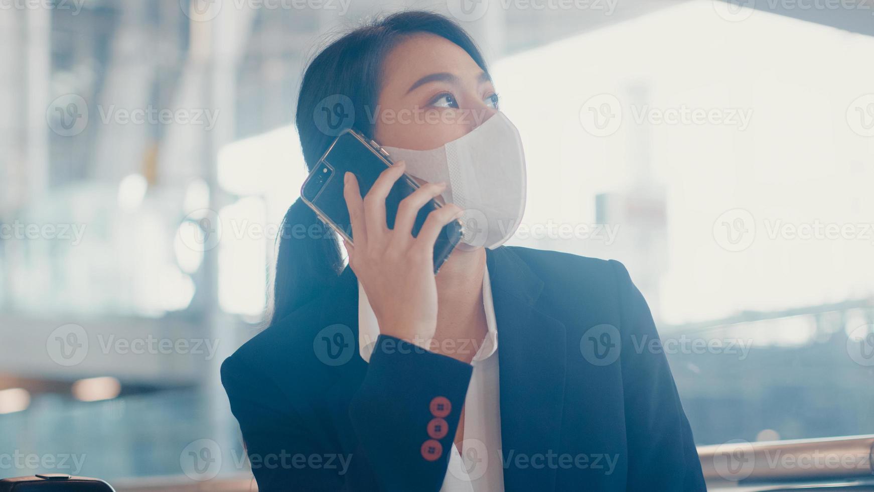 Une fille d'affaires asiatique intelligente porte un costume assis avec une valise sur un banc, appelez un téléphone intelligent avec un partenaire, attendez le vol à l'aéroport. navetteur de voyage d'affaires dans la pandémie de covid, concept de voyage d'affaires. photo