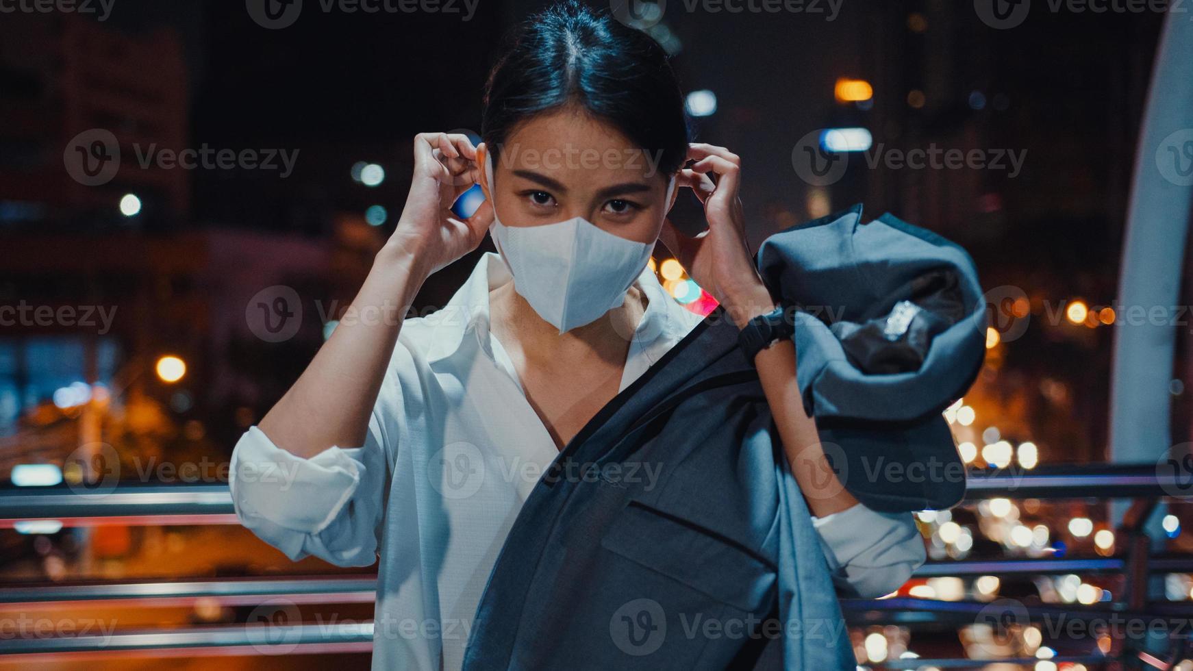 Une jeune femme d'affaires asiatique réussie dans des vêtements de bureau de mode porte un masque médical souriant et regardant la caméra tout en étant heureuse seule à l'extérieur dans la nuit de la ville moderne urbaine. concept d'entreprise en marche. photo