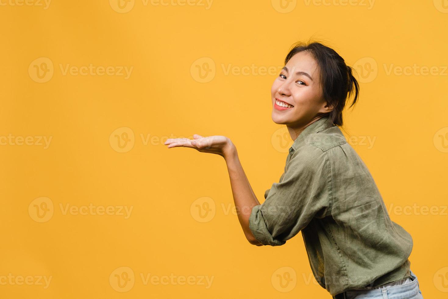 portrait d'une jeune femme asiatique souriante avec une expression joyeuse, montre quelque chose d'étonnant dans un espace vide dans un tissu décontracté et regardant la caméra isolée sur fond jaune. concept d'expression faciale. photo