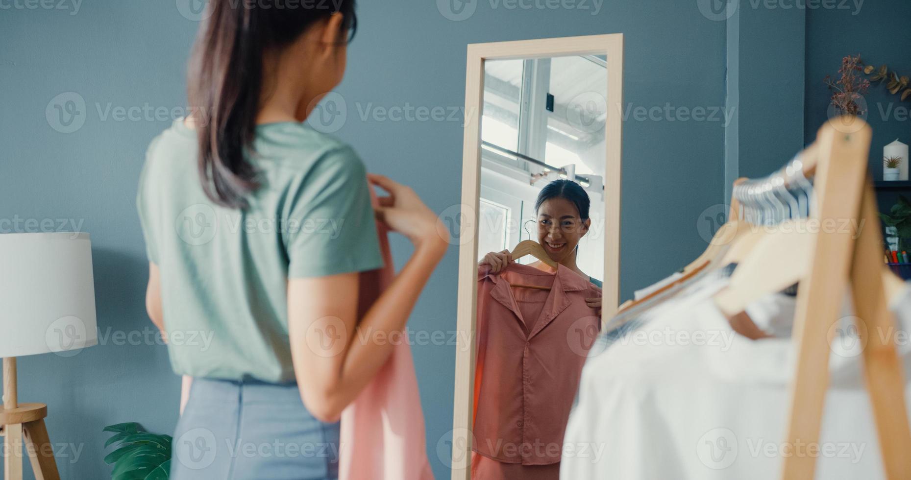 belle dame asiatique séduisante choisissant des vêtements sur un portemanteau s'habillant en se regardant dans un miroir dans le salon de la maison. fille pense quoi porter chemise décontractée. les femmes de style de vie se détendent à la maison concept. photo