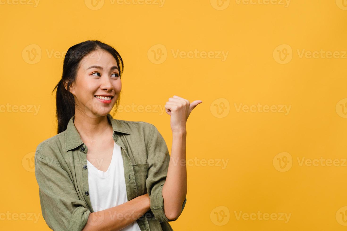 portrait d'une jeune femme asiatique souriante avec une expression joyeuse, montre quelque chose d'étonnant dans un espace vide dans des vêtements décontractés et debout isolé sur fond jaune. concept d'expression faciale. photo