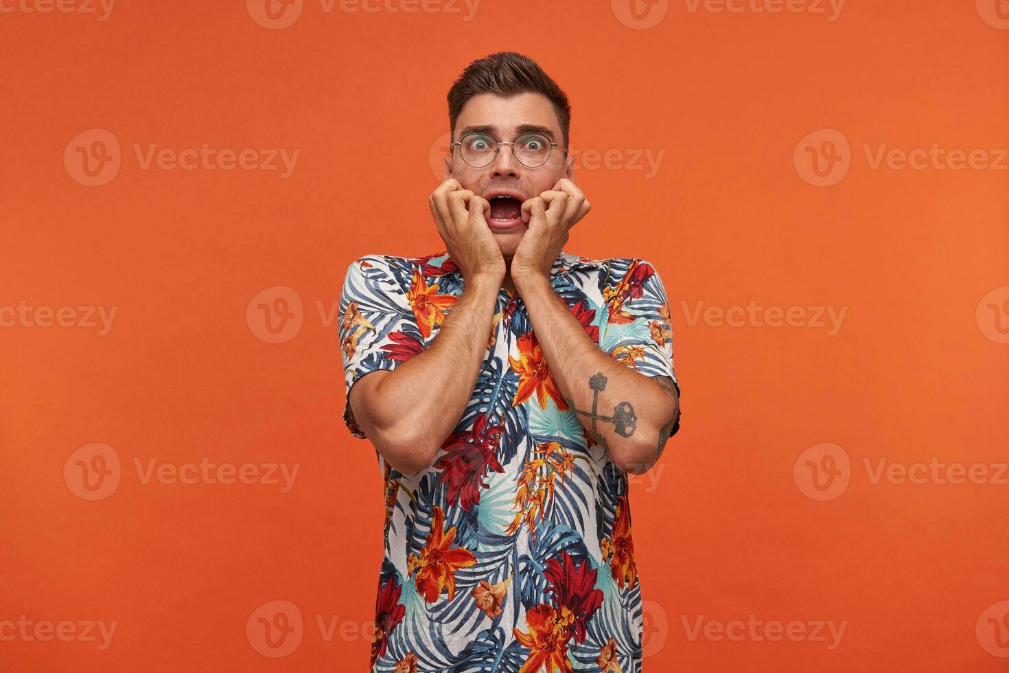 Jeune homme dans floral chemise avec le sien mains sur le visage et ouvert bouche des stands plus de le Orange arrière-plan, regards effrayé à caméra, portant des lunettes photo