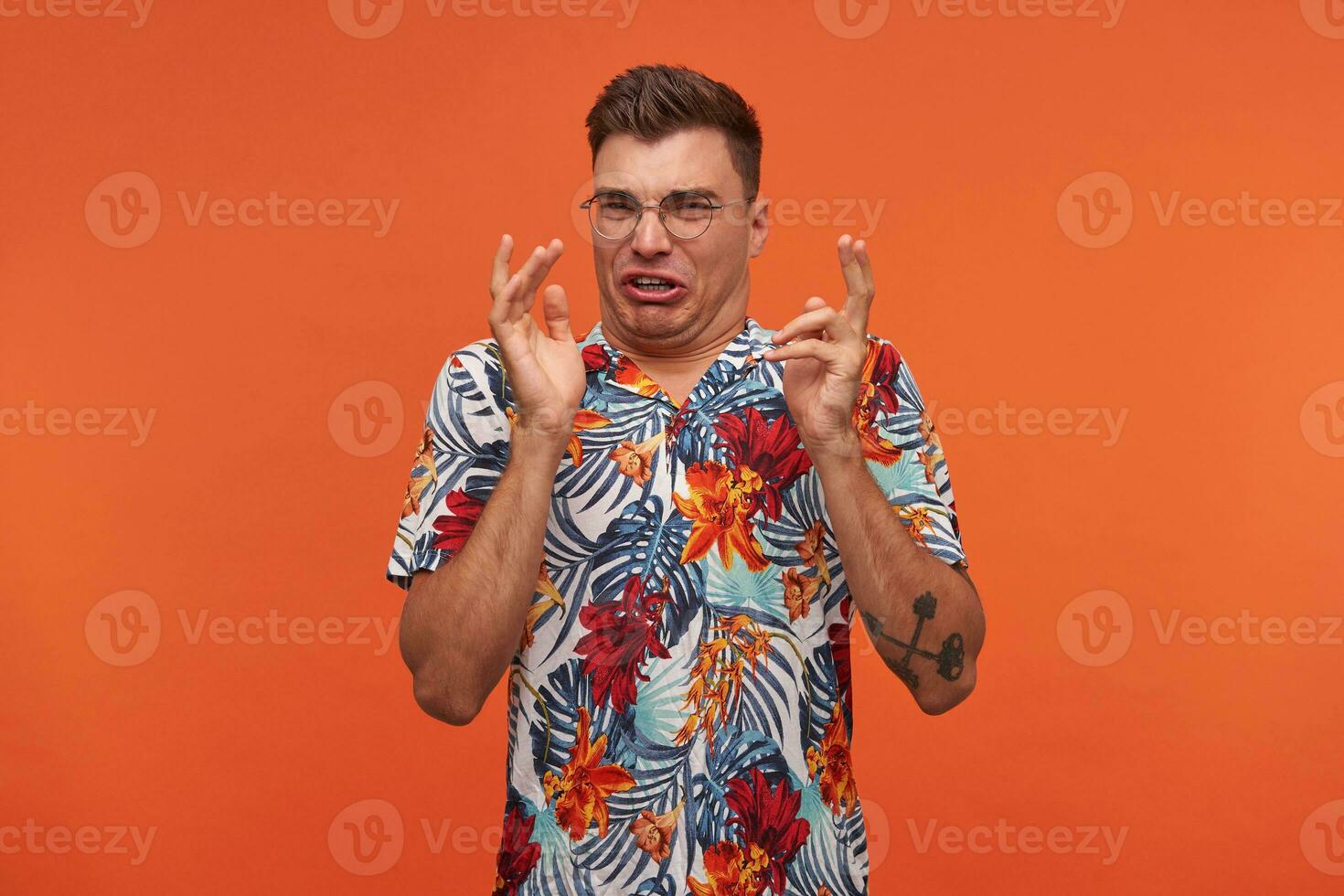 photo de Jeune cheveux courts gars avec élevé mains, montrant dégoûter, portant chemise avec floral impression et lunettes, fabrication grimace, permanent plus de Orange Contexte