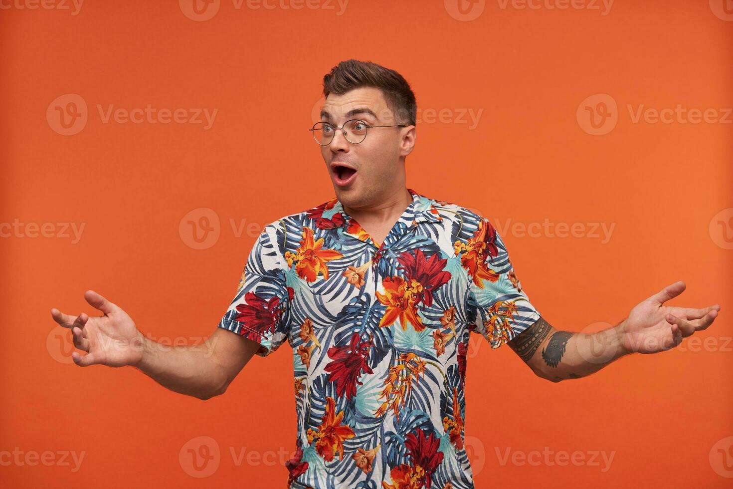 surpris Jeune homme dans floral chemise hausse les épaules avec ouvert bouche, permanent plus de Orange arrière-plan, portant lunettes, à la recherche une façon photo