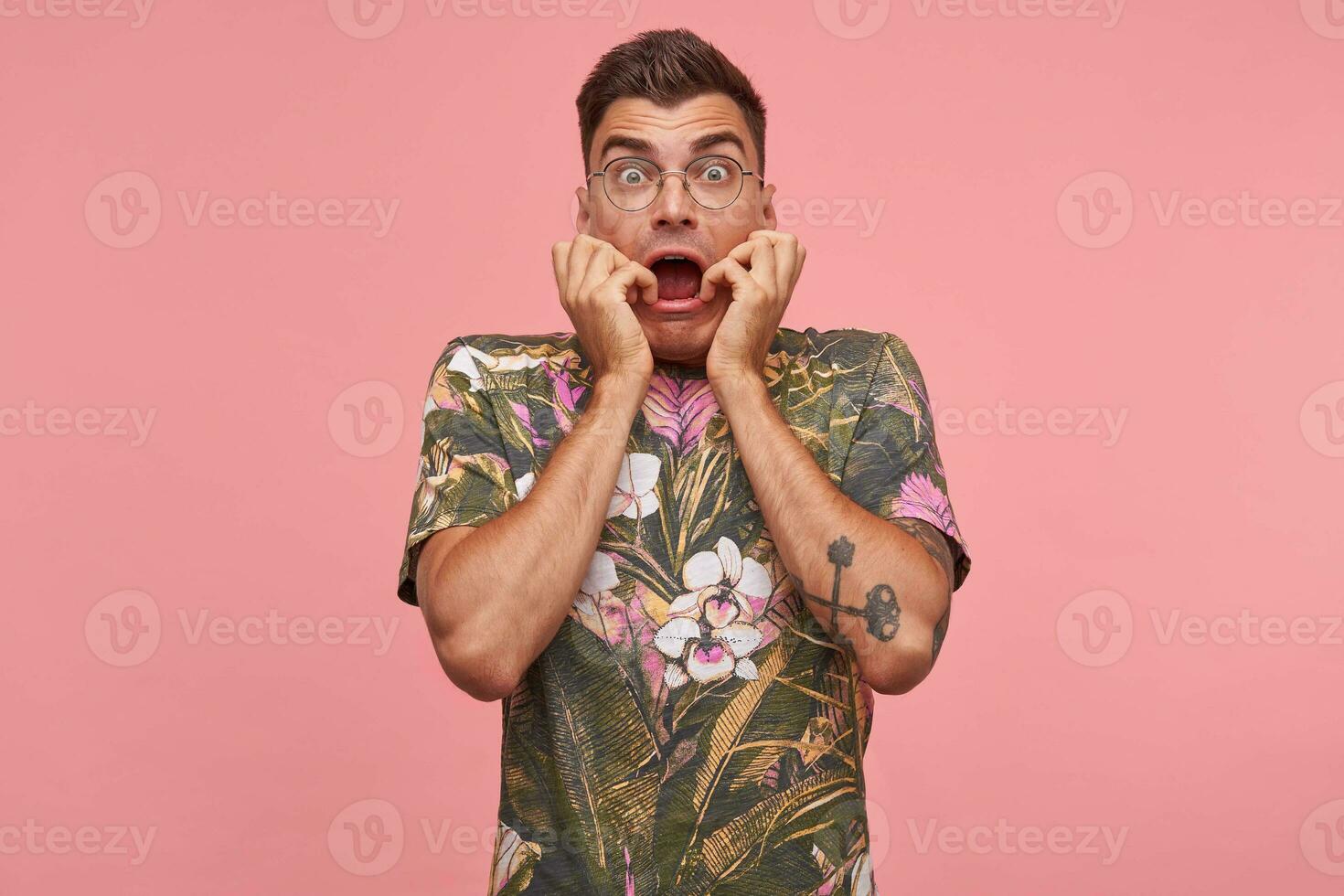 coup de une Jeune gars avec le sien mains sur le visage et ouvert bouche, permanent plus de le rose arrière-plan, regards effrayé à caméra, portant des lunettes et fleuri T-shirt photo