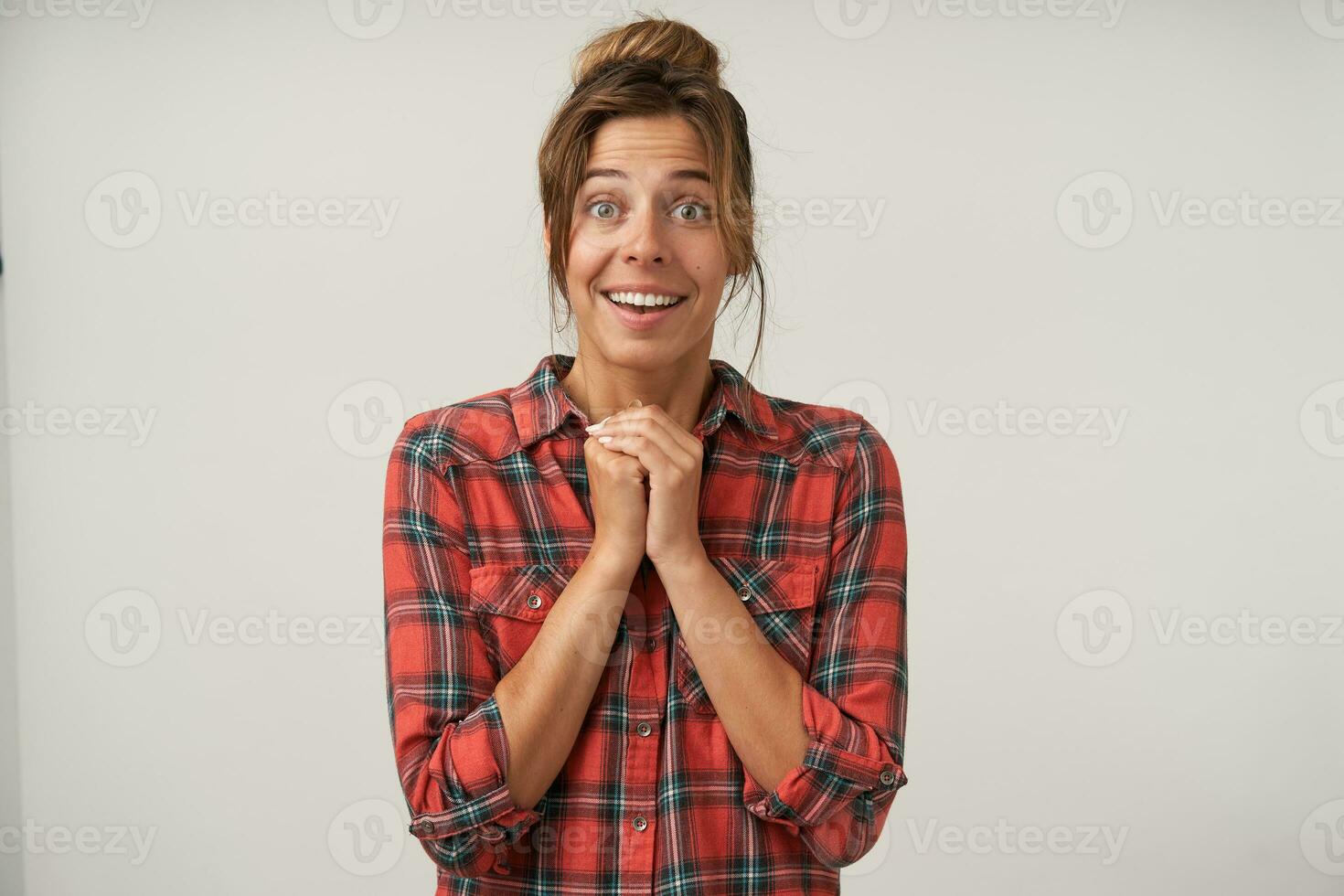 studio photo de Jeune marron aux cheveux Dame avec chignon coiffure arrondi avec surprise sa vert yeux tandis que à la recherche à caméra et en gardant mains plié, isolé plus de blanc Contexte