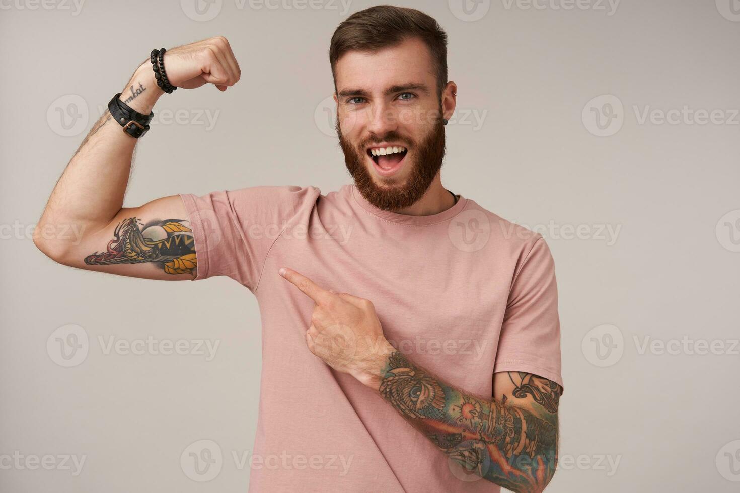 joyeux Jeune barbu brunette Masculin avec branché la Coupe de cheveux portant beige T-shirt posant plus de blanc arrière-plan, montrant sur le sien élevé main et sentiment fier à propos fort muscles photo