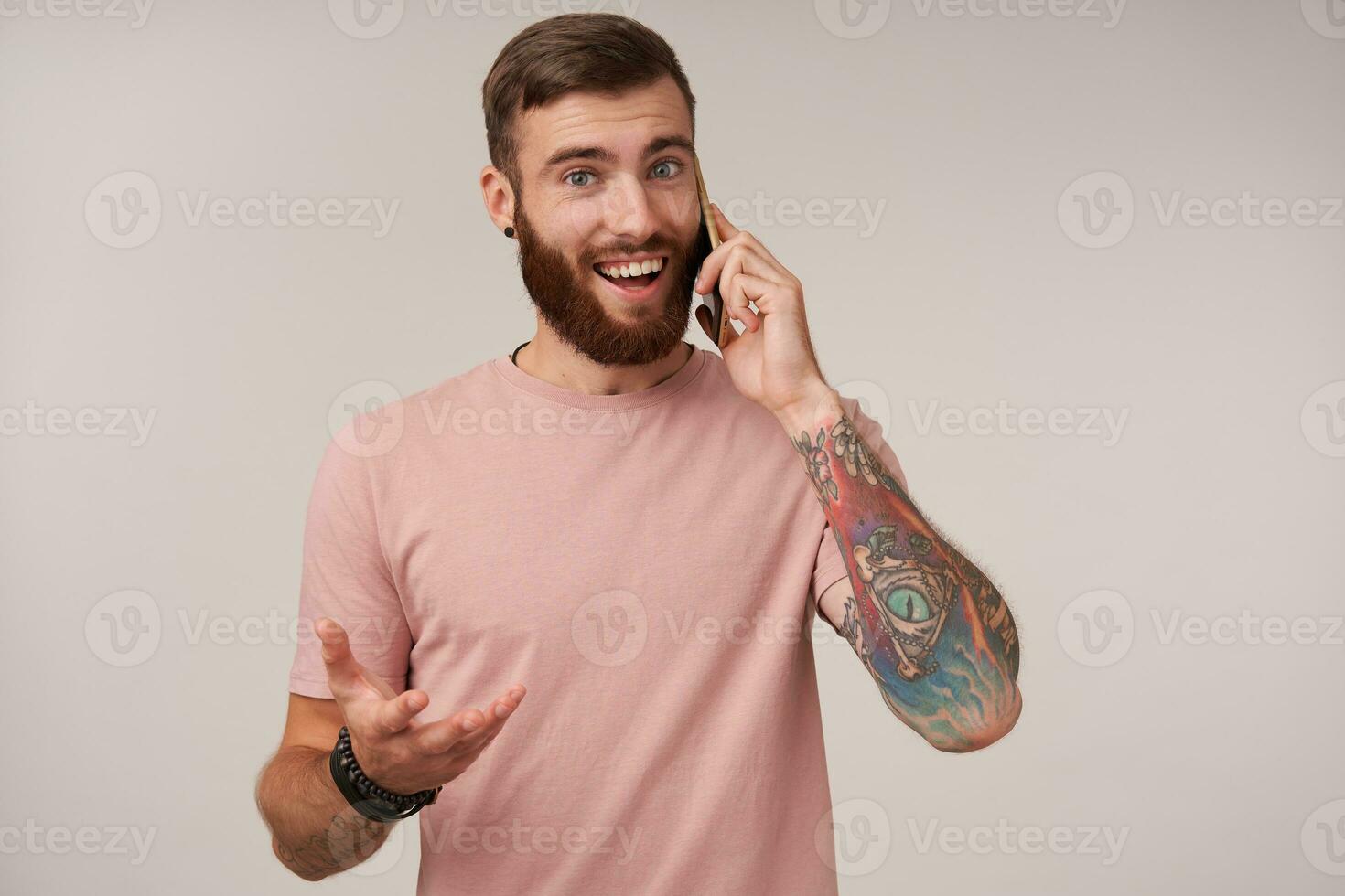 de bonne humeur barbu brunette gars avec court la Coupe de cheveux portant décontractée vêtements et branché accessoires tandis que permanent plus de blanc arrière-plan, ayant agréable conversation sur téléphone et à la recherche joyeusement à caméra photo