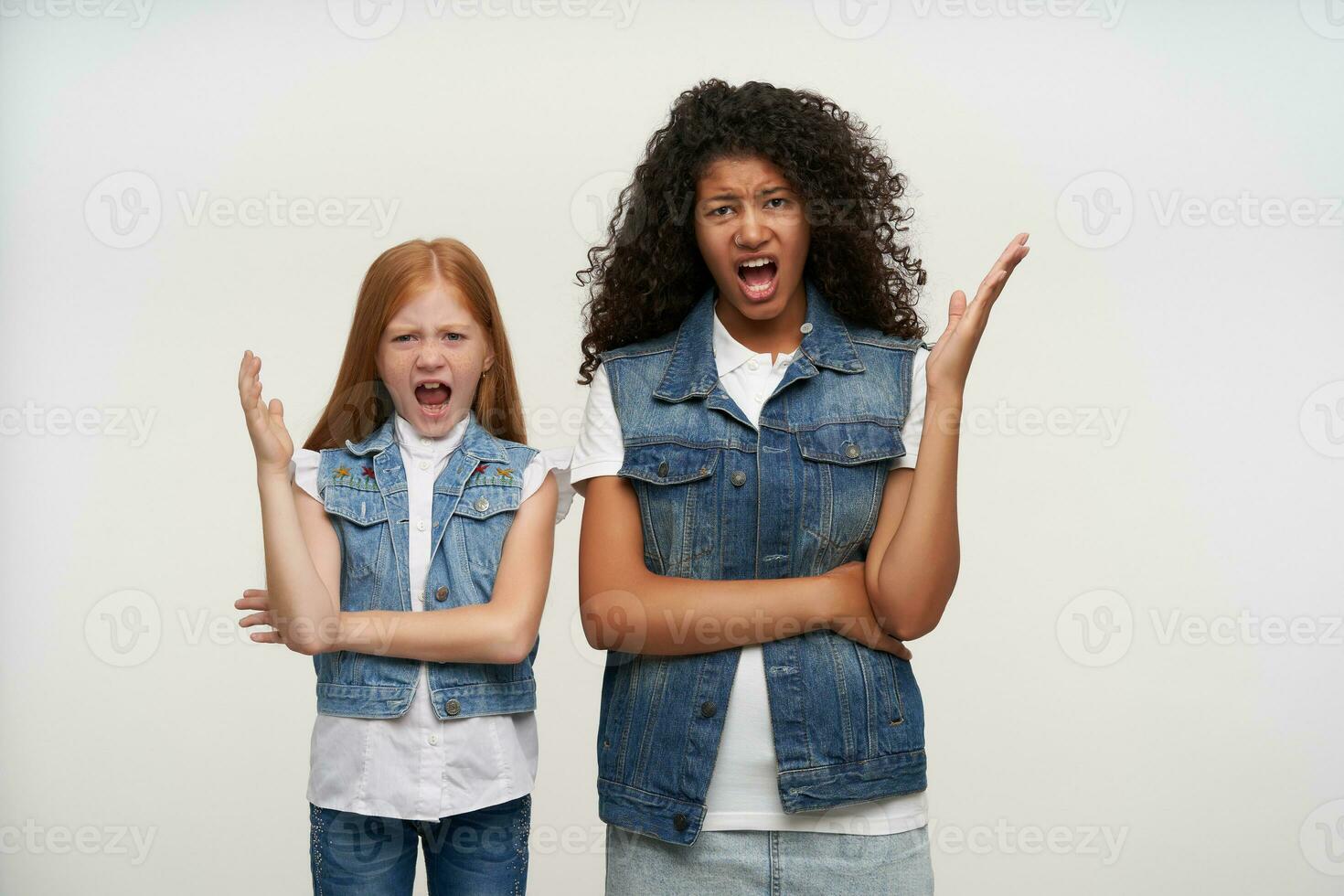 studio coup de irrité Jeune couple de frisé foncé aux cheveux femelle et rusé longue aux cheveux fille dans décontractée vêtements à la recherche avec colère à caméra et en criant agacé, isolé plus de blanc Contexte photo