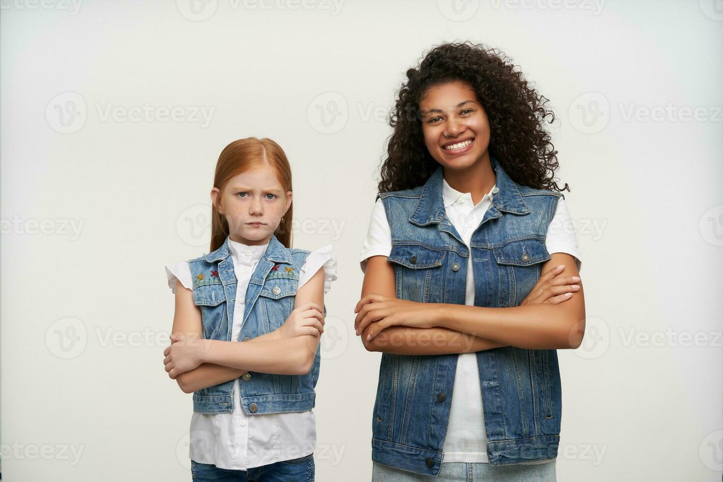 couple de Jeune attrayant Dames dans jeans gilets et blanc chemises montrant joie et tristesse tandis que permanent plus de blanc arrière-plan, en gardant mains plié sur leur coffres photo