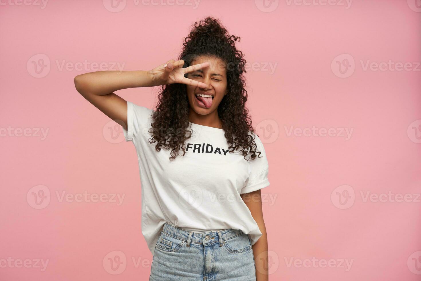 coup de marrant Jeune frisé longue aux cheveux brunette femme avec foncé peau fermeture un œil et montrant langue à caméra, pliant la victoire soupir et en gardant il près sa affronter, isolé plus de rose Contexte photo