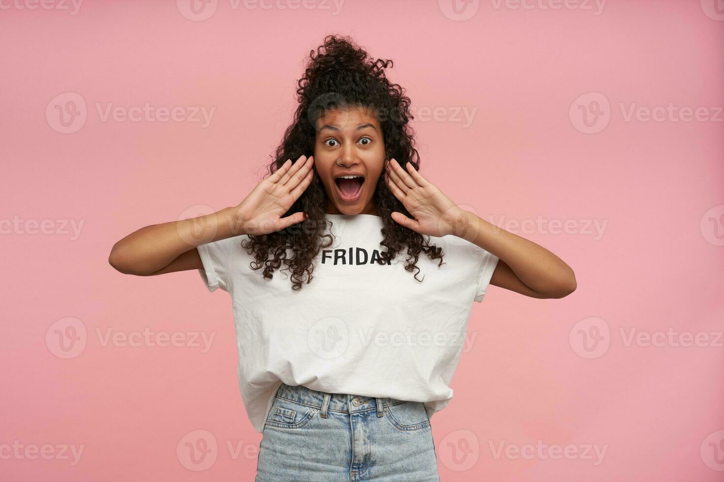 excité jolie Jeune frisé longue aux cheveux brunette femme avec foncé peau élevage paumes à sa bouche et à la recherche à caméra avec large yeux et bouche ouvert, isolé plus de rose Contexte photo