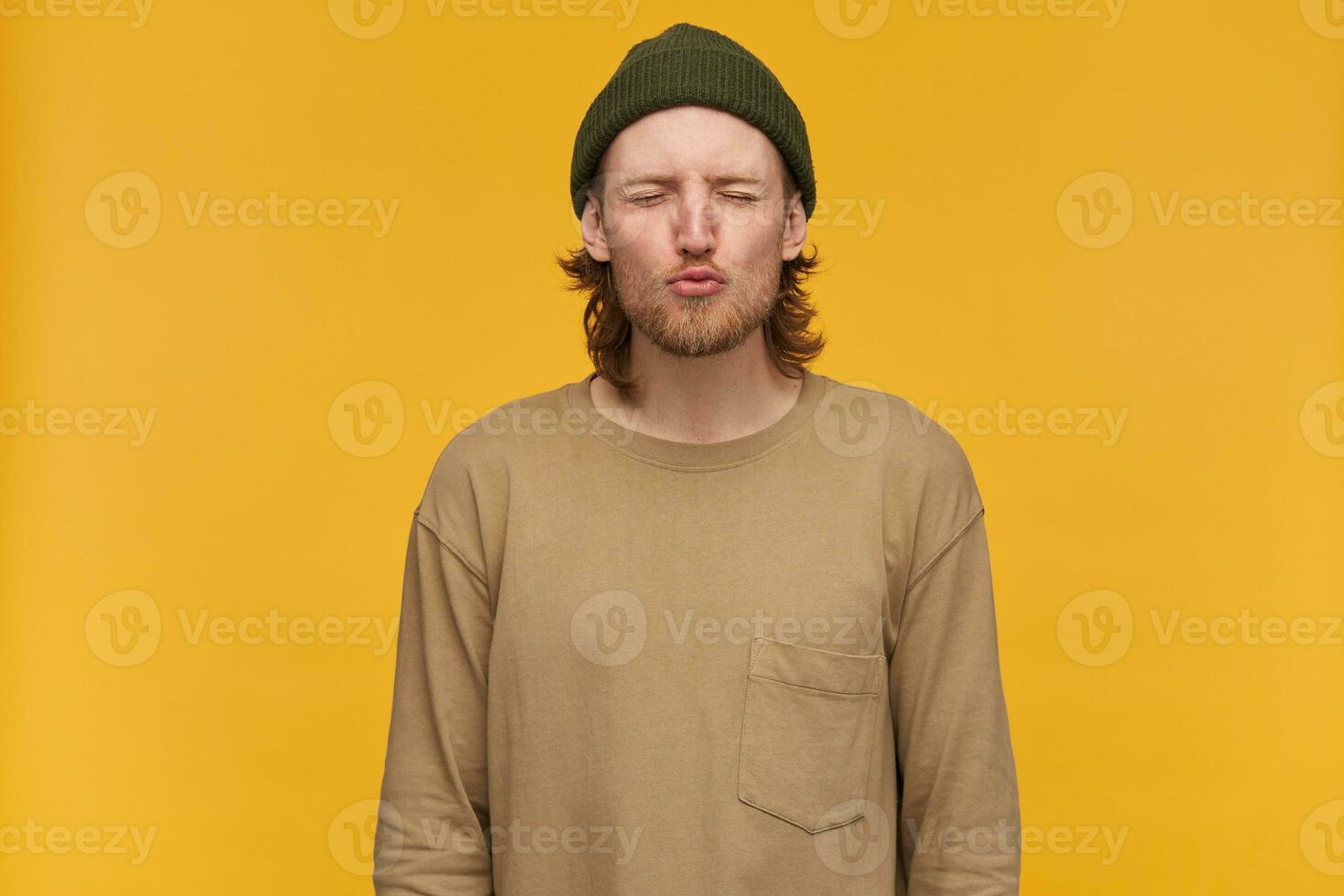 Jeune homme, cool gars avec blond cheveux, barbe et moustache. portant vert bonnet et beige chandail. sacs à main le sien lèvres dans une baiser et garde yeux fermé. supporter isolé plus de Jaune Contexte photo