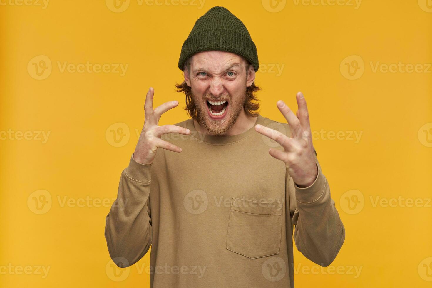 en colère à la recherche homme, mécontent barbu gars avec blond cheveux. portant vert bonnet et beige chandail. fait du effrayant grimace. en train de regarder à le caméra isolé plus de Jaune Contexte photo