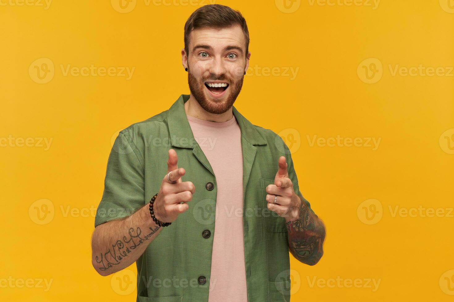 de bonne humeur attrayant barbu Jeune homme avec tatouage souriant et montrer du doigt sur vous à caméra par deux les doigts plus de Jaune Contexte photo